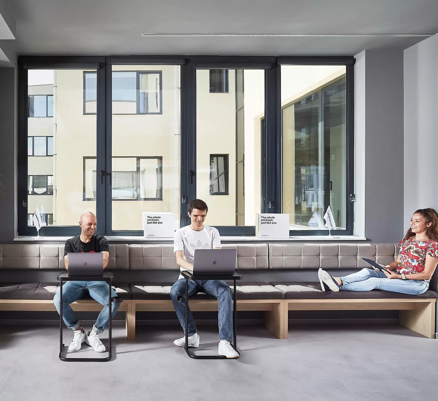 three persons working on their laptops in front of a large window in an office