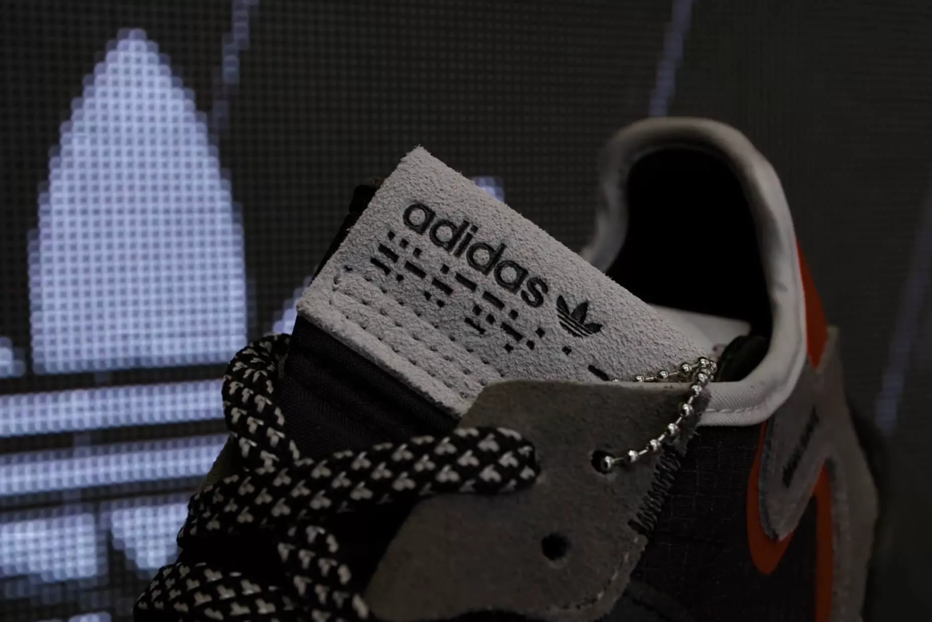 A close-up of an Adidas sneaker shows the brand’s logo on the tongue, featuring black and white laces and a mix of gray and orange details.